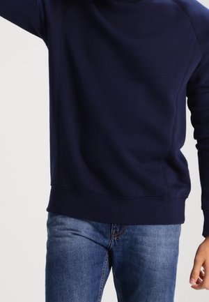 Person trägt einen marineblauen Sweatshirt mit langen Ärmeln und blaue Jeans, eine Hand ist erhoben und die andere entspannt seitlich.