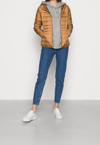 Bruine pufferjas met een capuchon, grijze hoodie eronder, blauwe skinny jeans en witte sneakers; gladde textuur en gewatteerd design.