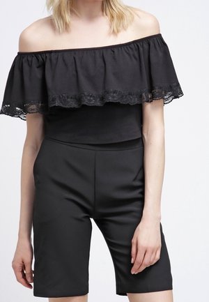 Femme portant un haut noir à épaules dénudées avec une bordure en dentelle et un short noir cintré taille haute arrivant au genou, sur fond uni.