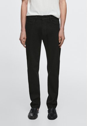 Sorte, lige bukser båret med sorte sko og en hvid skjorte, vist fra talje til fødder mod en ensfarvet baggrund.