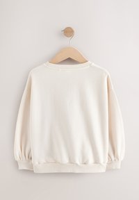 Bluza de culoare crem cu textură moale, guler rotunjit și manșete și tiv elasticizate. Atârnă pe un umeraș de lemn, pe un fundal neutru.