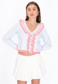 Ljusblå cardigan med rosa volangdetaljer, V-ringning och tre knappar. Materialet verkar mjukt. Buren över en veckad vit kjol.