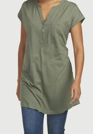 Kvinde iført olivengrøn kortærmet tunika med knappet V-hals og brystlomme kombineret med blå jeans mod en ensfarvet baggrund.
