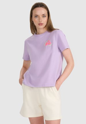Kobieta ubrana w lawendowy t-shirt z małą różowo-żółtą grafiką i kremowe szorty, stojąca z rękami w kieszeniach.