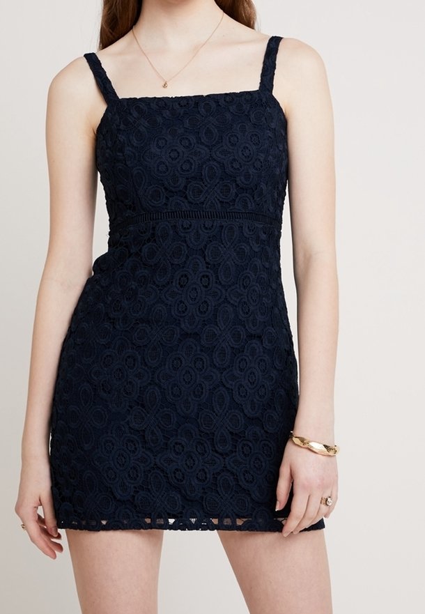 Robe en dentelle bleu marine sans manches avec encolure carrée, taille cintrée, portée avec un bracelet doré, une bague et un collier délicat.