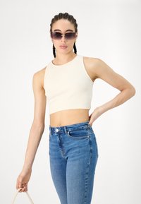 Femme aux cheveux tressés portant des lunettes de soleil surdimensionnées, un crop top beige sans manches et un jean bleu taille haute, tenant un sac beige.