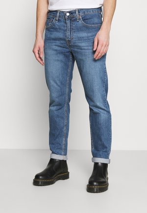 Homme portant un jean en denim bleu avec les revers et des bottines noires en cuir, sur un fond uni, les mains détendues le long du corps.