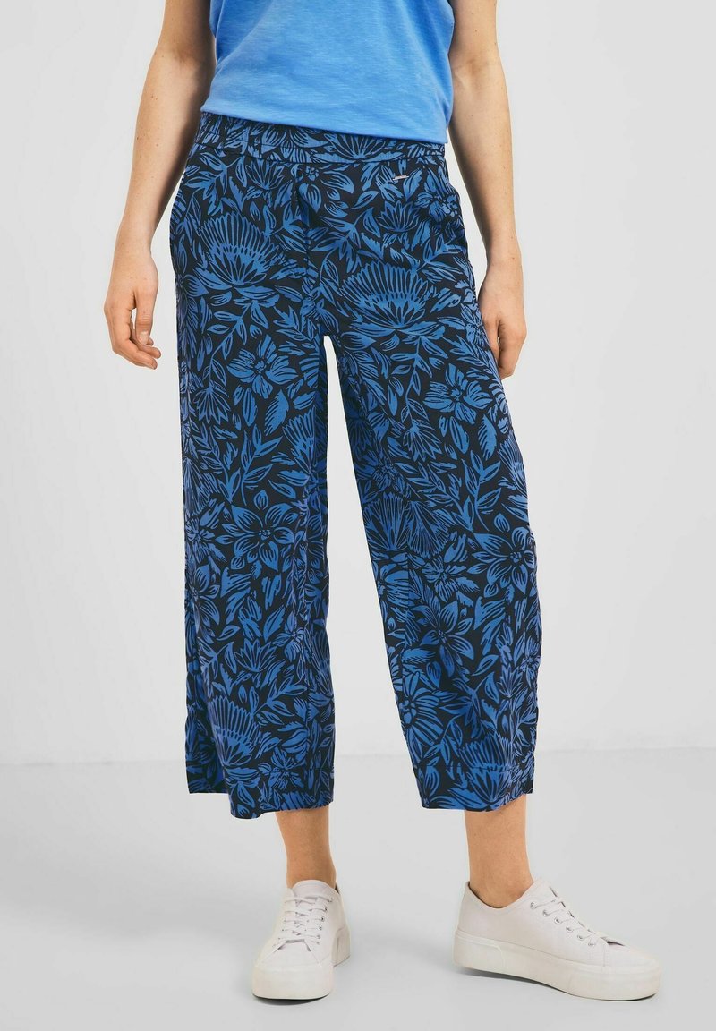 Femme portant un pantalon large court bleu à fleurs, des baskets blanches et un haut bleu, debout devant un fond uni.
