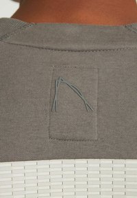 Grå sweatshirt med struktureret stof, et rektangulært mærke på ryggen og en diskret mønstret detalje i kanten.