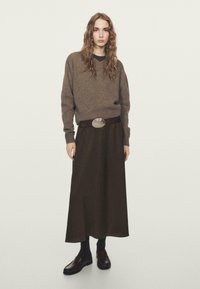 Femme aux longs cheveux ondulés portant un pull marron, une jupe midi marron foncé avec ceinture, des chaussettes noires et des mocassins marron, debout devant un fond uni.
