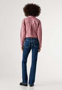 Sarkani un balti svītraina pogplūkšanas krekls ar garām piedurknēm, saskaņots ar tumši ziliem zvana pieguļošiem džinsiem, skatīts no aizmugures. Redzamas melnas kurpes.