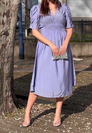 Kobieta ubrana w lawendową sukienkę midi i srebrne szpilki, trzymająca małą srebrną kopertówkę, stojąca na zewnątrz obok drzewa i balustrady.