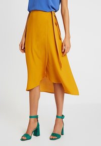 Gonna avvolgente gialla con orlo asimmetrico, realizzata in tessuto leggero. Abbinata a sandali con tacco a blocco color turchese.