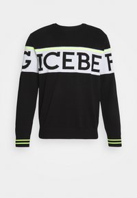 Pull noir à manches longues avec une bande horizontale blanche sur la poitrine et les manches, comportant de grandes lettres noires et des rayures jaune néon sur les poignets.