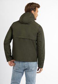 Oliven tonet, vandafvisende jakke med hætte, der har en rygspids og glat tekstur. Bæres med lyseblå jeans.