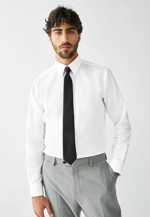 Man met krullend haar en baard, gekleed in een wit overhemd, zwarte stropdas en grijze broek, staand met één hand in de zak.