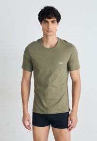 Olivegrünes Baumwoll-T-Shirt mit rundem Halsausschnitt und kurzen Ärmeln, mit einem weißen "BOSS"-Logo auf der Brust. Getragen mit schwarzen Shorts.