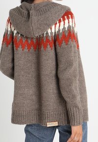 Sweter z dzianiny z kapturem, o brązowym ciele, z czerwono-białymi wzorami w zygzaki na ramionach oraz metką u dołu.