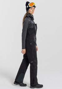 Black waterproof ski overall with adjustable straps, side pockets, reinforced knees, and a smooth texture. Worn with a dark top and ski goggles.