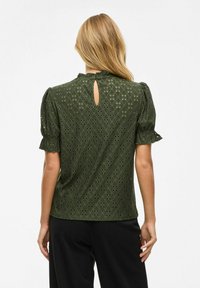 Groene blouse met opgeblazen mouwen, kantdetails en een keyhole opening op de rug. De stof heeft een geometrisch patroon en een gestructureerde afwerking.