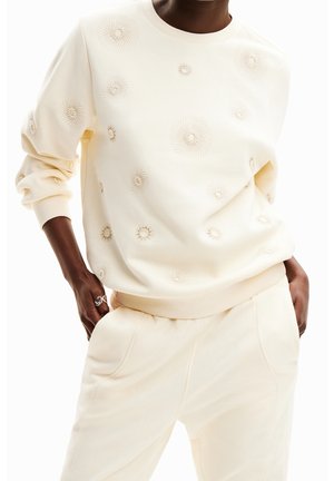 Person trägt cremefarbenen Pullover und Hose mit spiralförmiger Blumenstickerei, Hände in den Taschen und silberne Ringe an der linken Hand.