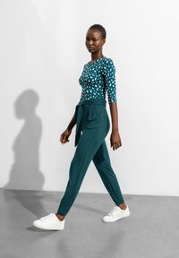 Tealfärgad långärmad skjorta med oregelbundna vita mönster, kombinerad med högmidjade tealfärgade byxor och vita sneakers. Modell går på grått golv.