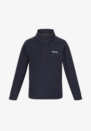 Garçon debout regardant sur le côté, portant une polaire zippée bleu marine, un pantalon cargo et des baskets sur un fond uni.