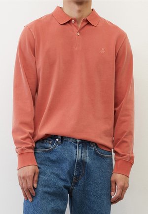 Homme portant un polo rouge délavé à manches longues avec deux boutons, associé à un jean en denim bleu, debout devant un fond uni.
