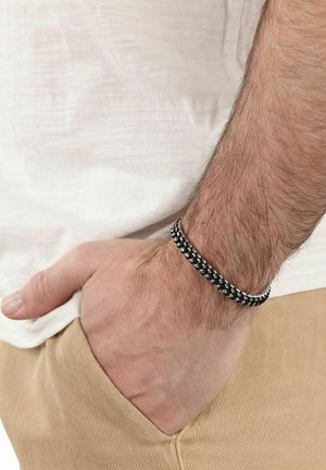 Polso maschile con braccialetto intrecciato nero e argento, mano parzialmente nella tasca dei pantaloni beige e che indossa una camicia bianca.