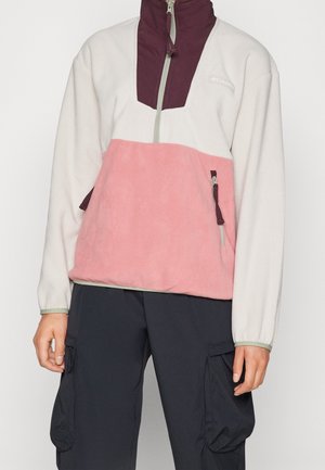 Osoba nosząca polarowy sweter color-block w kolorach białym, różowym i bordowym z półzamkiem z przodu oraz czarne spodnie bojówki z dużymi kieszeniami.
