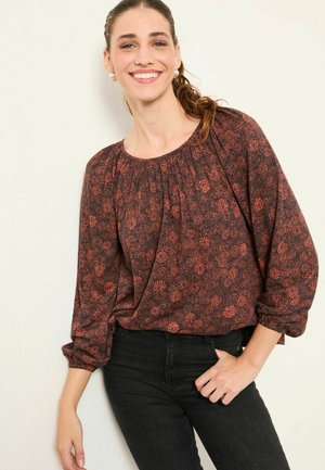 Langärmlige, dunkelrote Bluse mit Blumendruck, gerafftem Ausschnitt und lockerer Passform, kombiniert mit schwarzen Jeans. Glattes Material, dezenter Glanz.