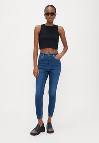 Γυναίκα με πλεξούδες, που φορά γυαλιά ηλίου, μαύρο αμάνικο κοντό τοπ, τζιν skinny μπλε ψηλόμεσο και μαύρα loafers με πάτο πλατφόρμα.