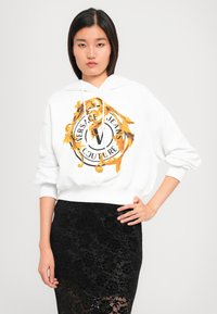 Giovane donna che indossa una felpa bianca con il logo dorato e nero di Versace Jeans Couture e una gonna in pizzo nero, in piedi con una mano sul fianco.