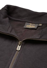 Schwarze Zip-Hoodie mit einem weichen Innenfutter, ausgestattet mit einem Metallreißverschluss und einem Etikett mit der Aufschrift "Switcher made with respect".