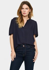 Marinblus med kort ärm och V-ringning, lös passform och textilstruktur, kombinerad med mörka jeans i denim med synlig sömnad.