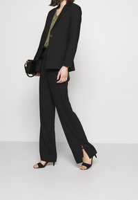 Zwarte blazer en set met wijde broek, voorzien van een knoopsluiting, strak stiksel en zijsplitten. Vergezeld van zwarte hoge hak sandalen.