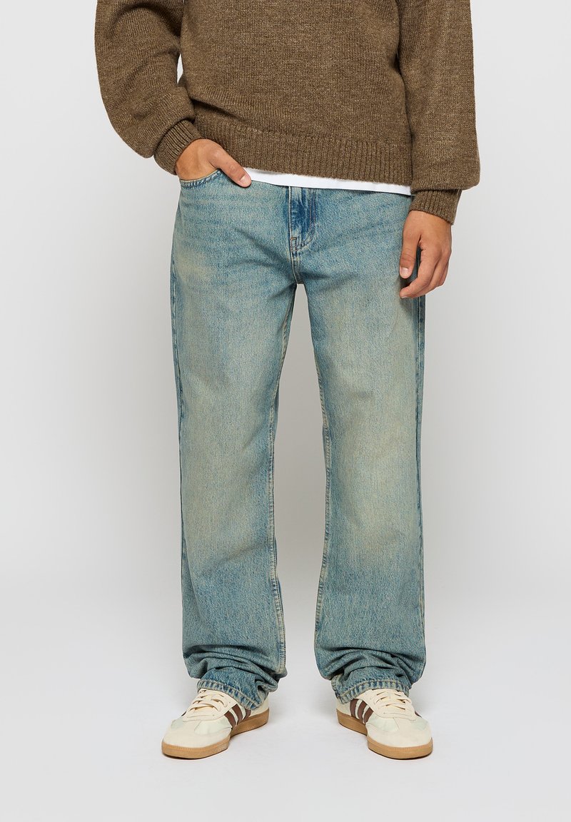 Lyseblå denimjeans med rett snitt, preget av en falmet vask, og iført en brun strikkegenser og kremfargede sneakers.
