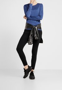 Camiseta deportiva azul de manga larga con escote redondo, combinada con mallas ajustadas negras. Una chaqueta negra brillante está atada a la cintura. Se llevan zapatillas negras.