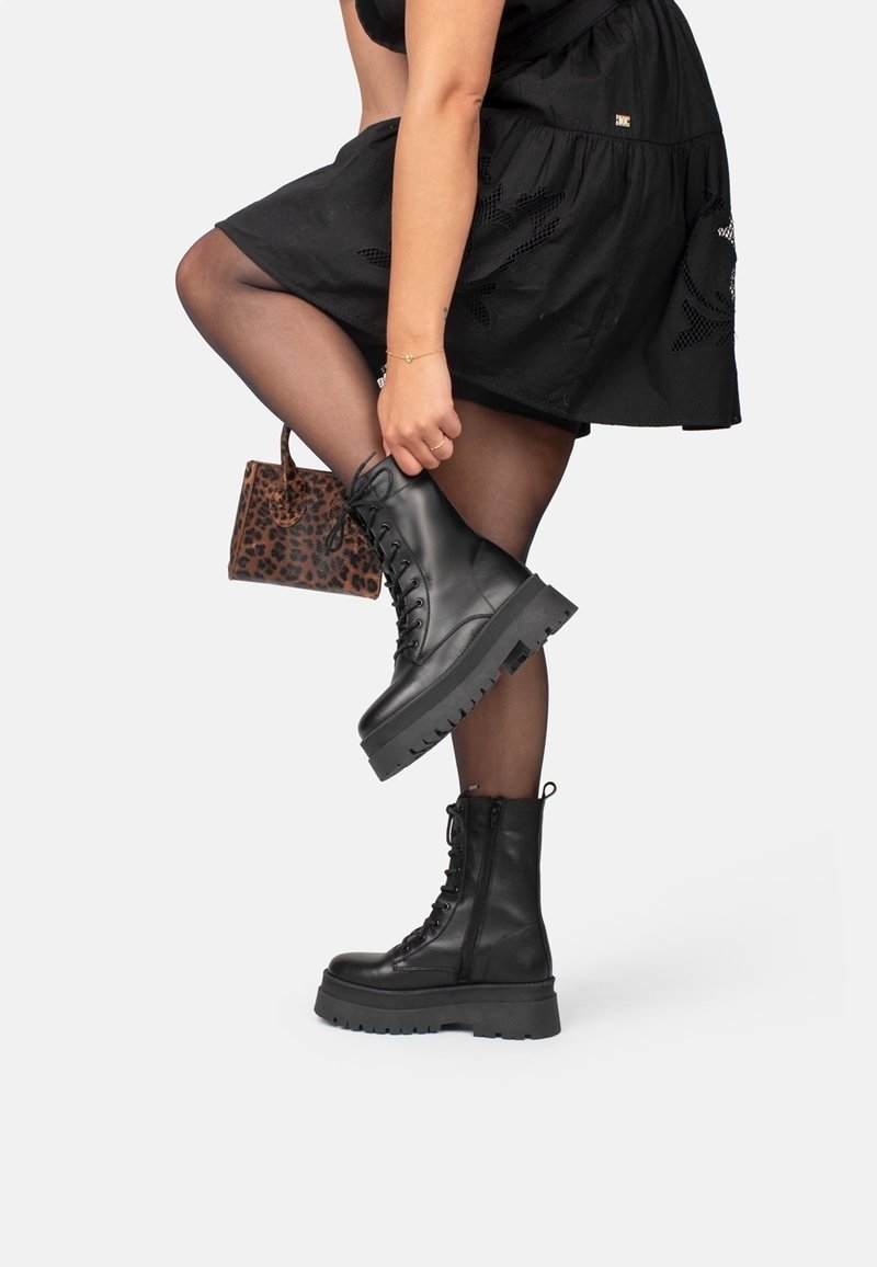 Personne portant des collants noirs transparents et une jupe noire lève une jambe, tenant une cheville avec des bottes de combat noires à lacets et un sac imprimé léopard à proximité.