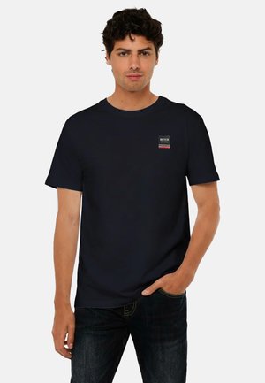 Giovane uomo con capelli ricci scuri che indossa una maglietta nera a maniche corte con un piccolo logo sul petto, in piedi con una mano in tasca.