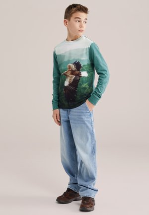 Rövid hajú fiú áll, türkiz hosszú ujjú pólót visel repülő sas mintával, világoskék farmert és barna tornacipőt, oldalra néz.