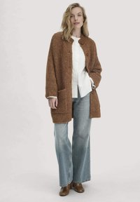 Frau in einem braunen Strickcardigan, weißem Hemd, weit geschnittenen Jeans und braunen Stiefeletten, die mit einer Hand in der Tasche steht.