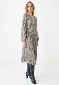 Jongeman staat, gekleed in een lange mouw panterprint jurk met een strik om de taille en zwarte kniehoge laarzen, op een effen witte achtergrond.