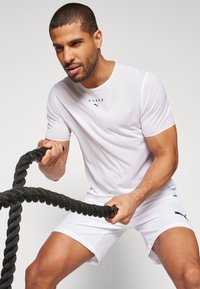 T-shirt blanc à manches courtes en tissu texturé avec un logo noir, associé à un short blanc, tenant une épaisse corde de combat noire.
