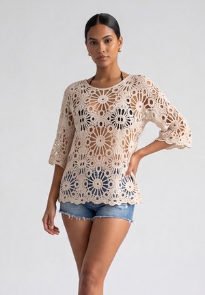 Vrouw met een beige gehaakte top over een zwarte bikini en spijkershorts, staand met één hand op de heup tegen een effen achtergrond.