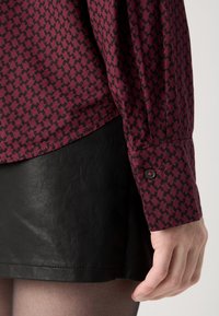 Camisa de botones color marrón con un patrón de pata de gallo, combinada con una falda corta de cuero negro texturizado. Detalles de botones visibles en las mangas.