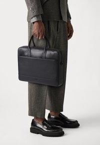 Svart skinnportefølje med doble håndtak, glatt finish og enkel design. Har en frontlomme og glidelås.