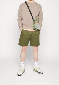 Brauner Sweatshirt, grüne Shorts und helle Turnschuhe. Eine kleine grüne Tasche mit grauem Netz und orangen Akzenten. Weiße Socken sichtbar.