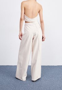 Kvinna som står på en blå matta iförd ljusbeige vida byxor och en matchande rygglös cropped top, vänd bort från kameran.