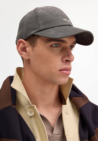 Young man wearing a dark gray baseball cap, beige coat with popped collar, and brown sweater, looking to the right on a light background.